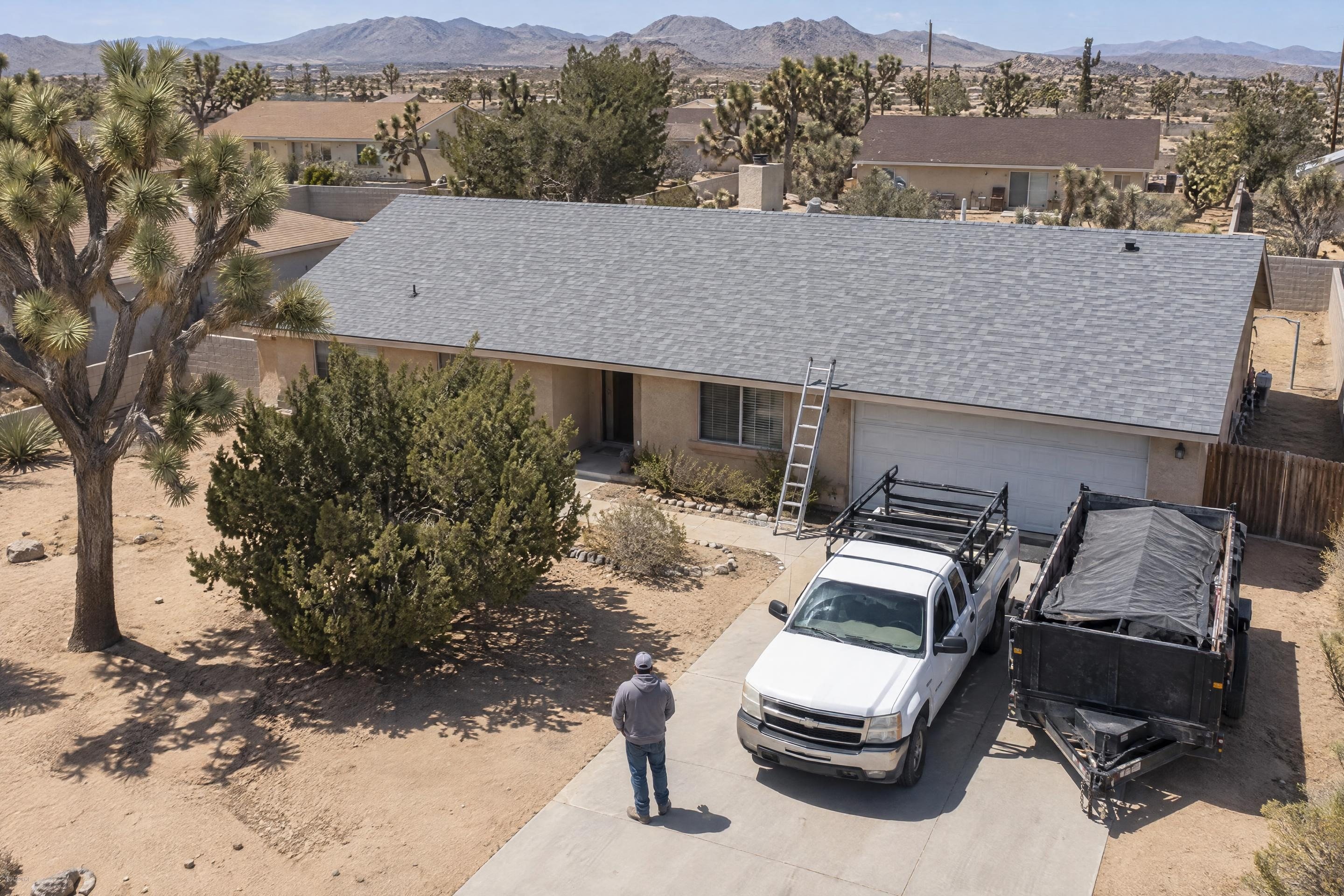 A work truck and dump trailer in a driveway under a new light grey shingle roof, seen from a drone